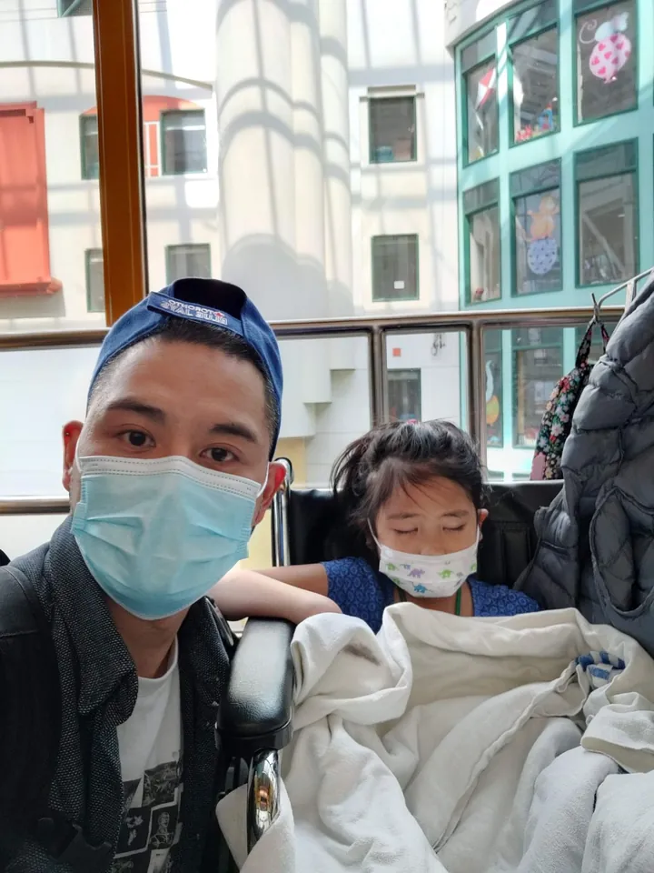 A selfie in the hospital atrium: me in a mask and ball cap, my daughter asleep under a blanket in a wheelchair.