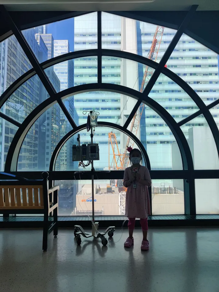 My daughter, in a face mask, stands beside her IV pole at a tall arched hospital window overlooking the city.