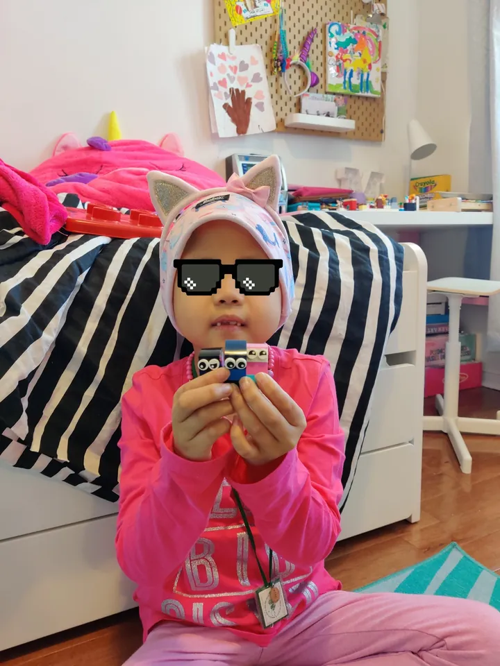 My daughter, in a cat-ear headband and pixel-art sunglasses, holds up three LEGO Ninja-Bloks.